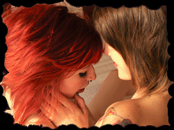 A girl is lying back, topless. Another girl is pulling down her shorts, exposing blue panties while her mouth is poised to kiss the skin.